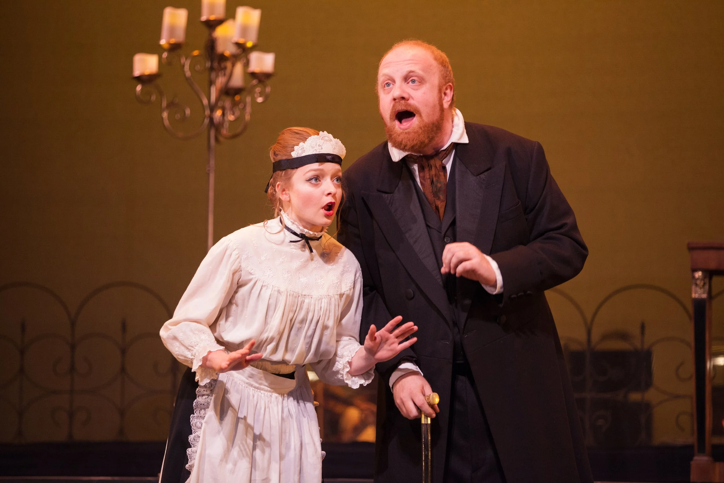 Two actors in period costumes performing on stage, a woman with red hair and a man with a beard, both looking surprised or emotional.