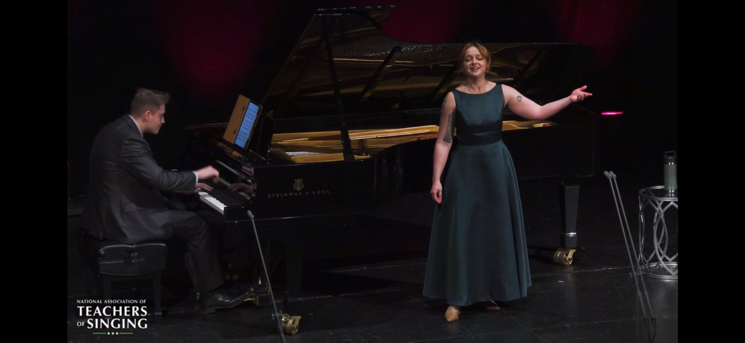 A woman in a long dark green dress singing on stage with a pianist playing a grand piano.