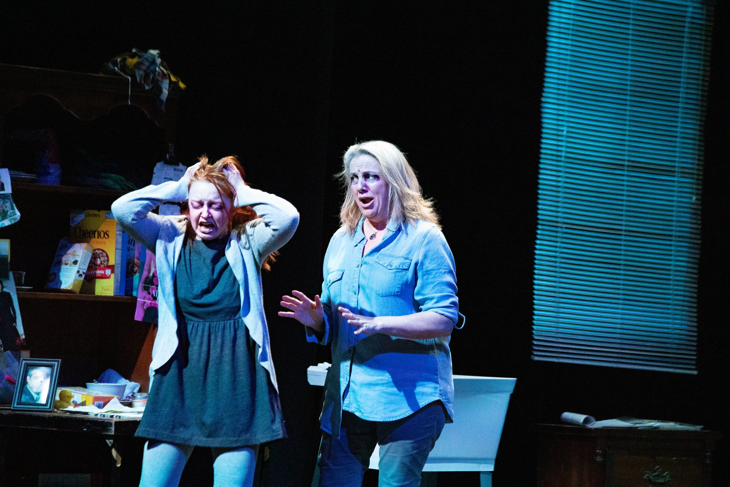 In a dramatic opera scene, one young woman has her hands in her hair with a pained expression on her face. Another woman attempts to comfort her.