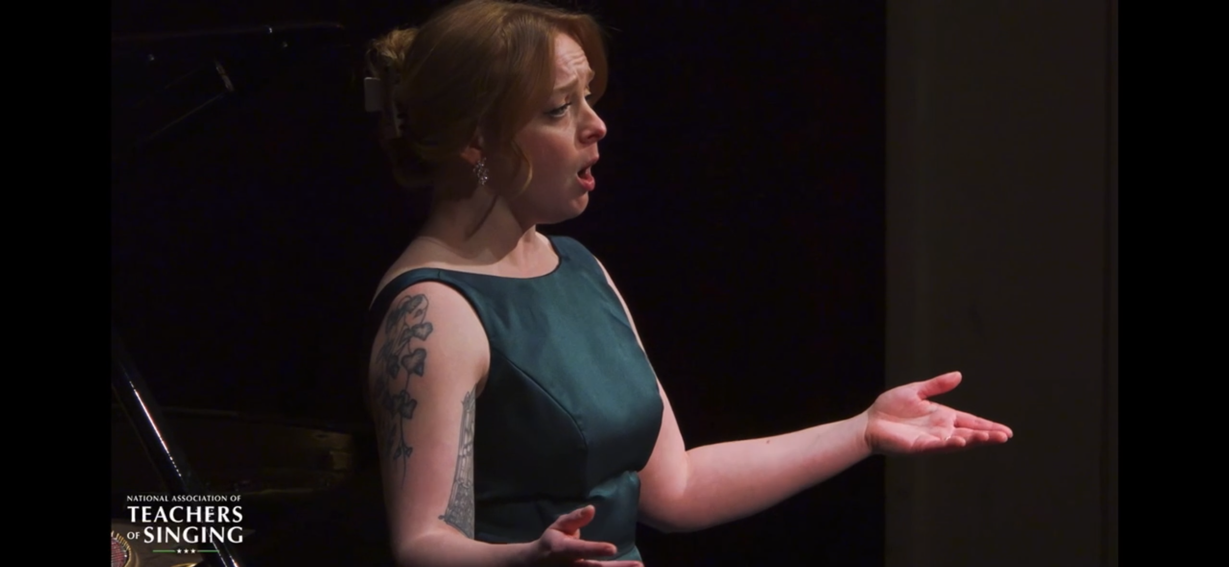 A woman singing with her eyes closed and hand raised, wearing a teal dress, with tattoos on her left arm, and short red hair, on a dark stage.