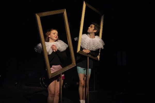 Two young performers stand behind large empty picture frames on sticks, engaging in a theatrical act on stage with a dark background.