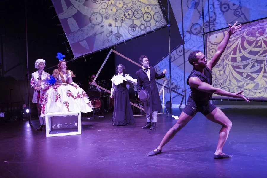 Ballet performers on stage, with one dancer in front striking a pose, and four actors in period costumes behind, with musical instruments and large colorful gears projected on the background.