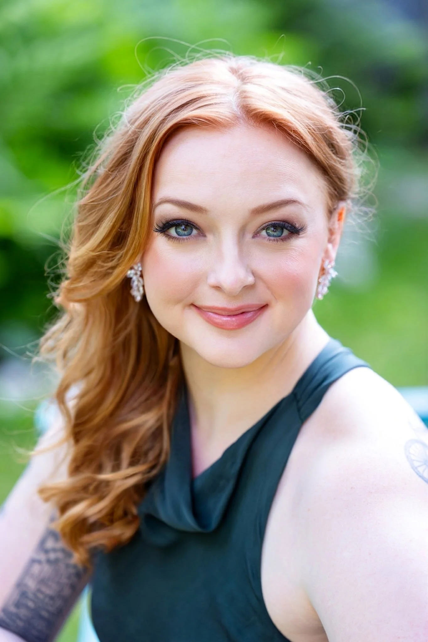 Close-up portrait of a woman with long red hair, blue eyes, and earrings, wearing a black sleeveless top, standing outdoors with green foliage in the background.