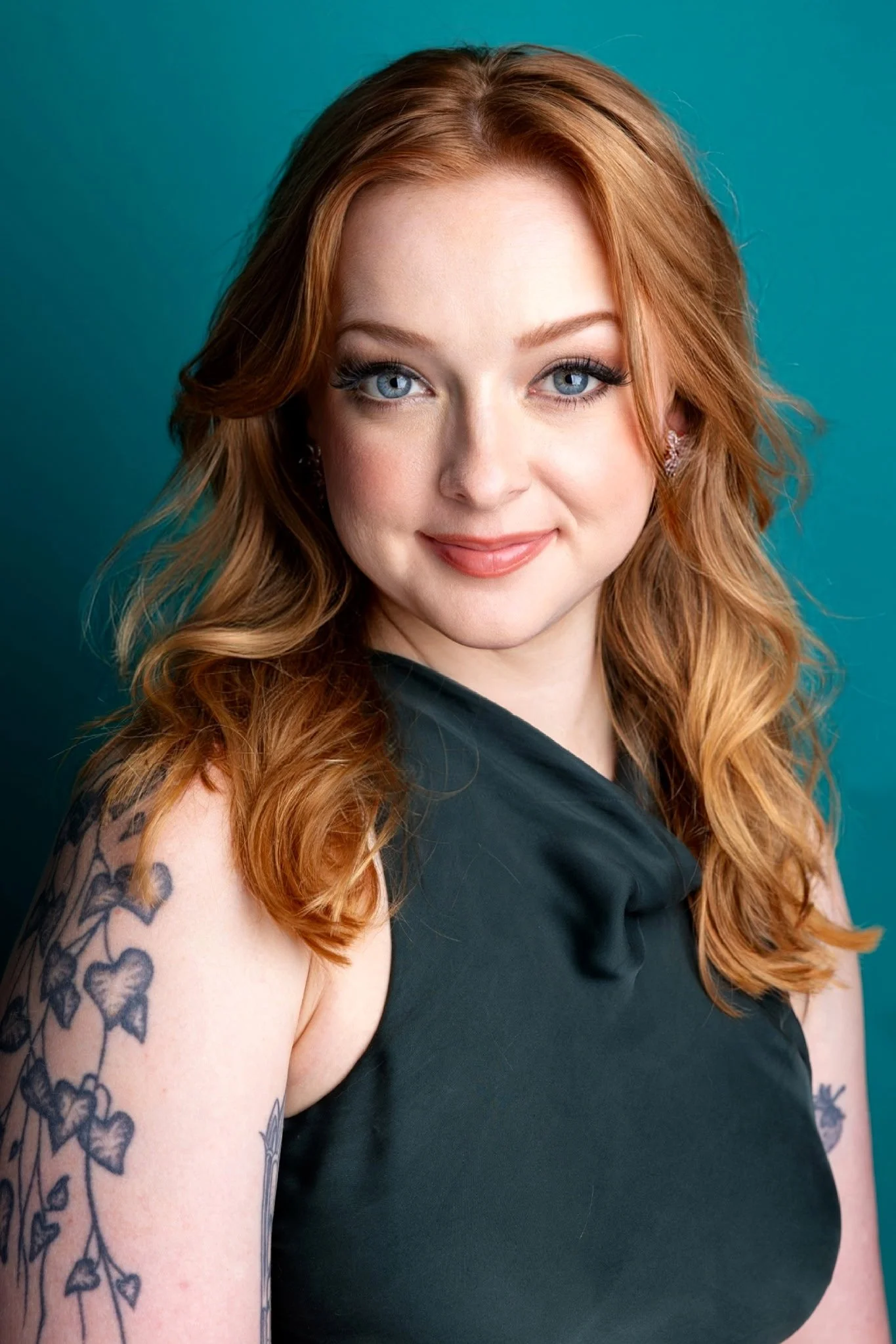 Close-up portrait of a woman with long red hair, blue eyes, and tattoos on her shoulder, wearing a black top, against a teal background.