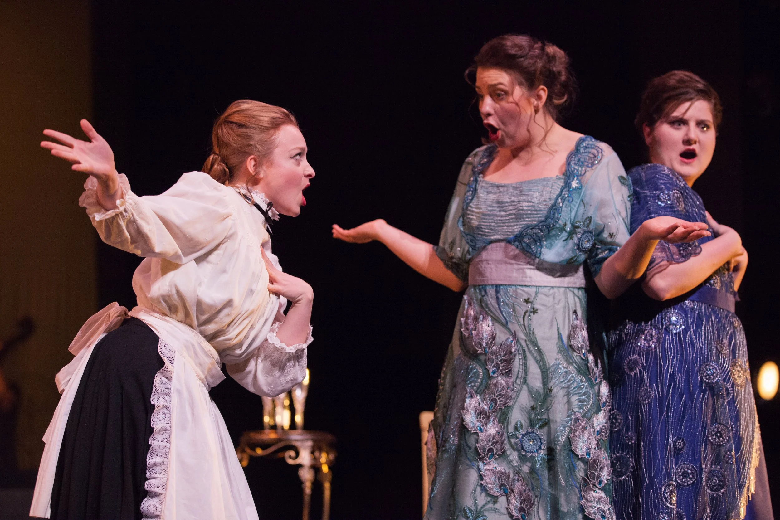 Three women dressed in period costumes performing in a theatrical play on stage, with one woman on the left passionately speaking or singing, and the other two women reacting with surprise or shock.