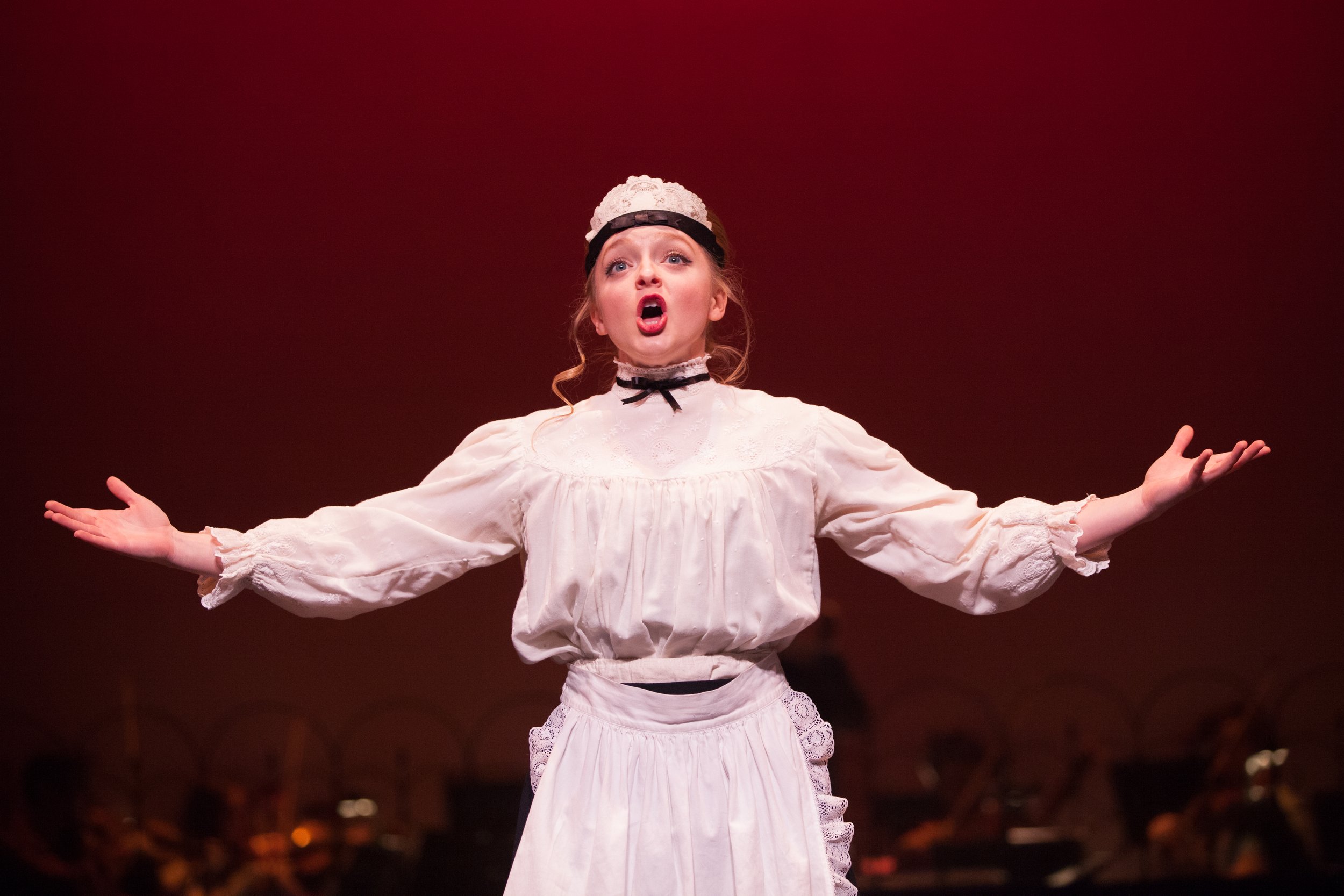 A young woman performing on stage, wearing a historical costume with a white lace headpiece, a high-neck blouse, and a long skirt, with arms outstretched and an expressive singing face against a dark red background.