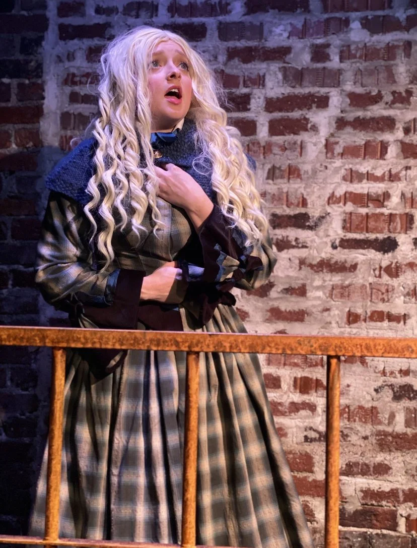 A woman with long, curly blonde hair wearing a plaid dress and dark coat, standing behind a rusted metal railing against an exposed brick wall, looking distressed and holding her hand to her chest.