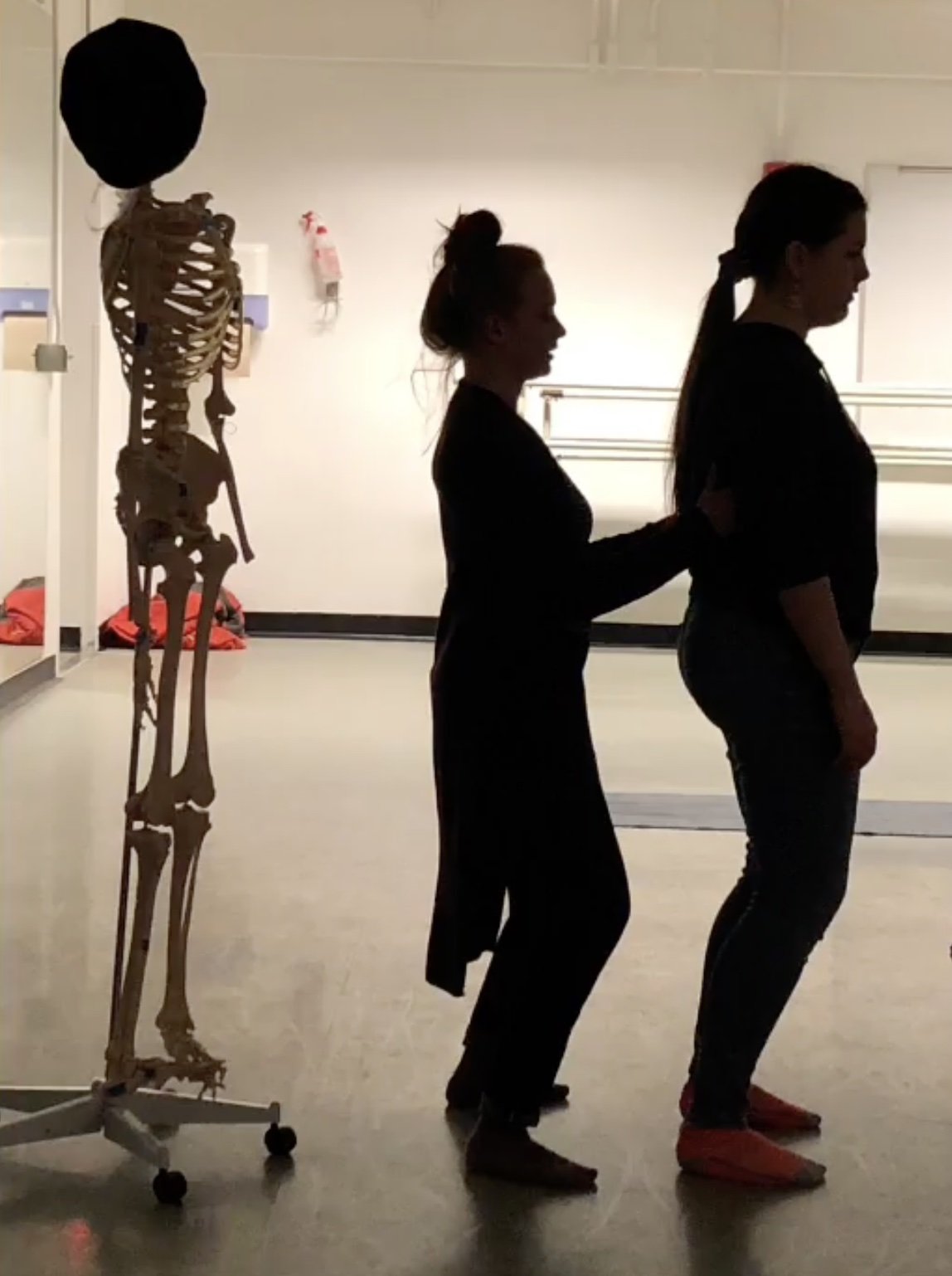 In front of a skeleton model, a young woman adjusts the posture of another young woman during an Alexander Technique session.