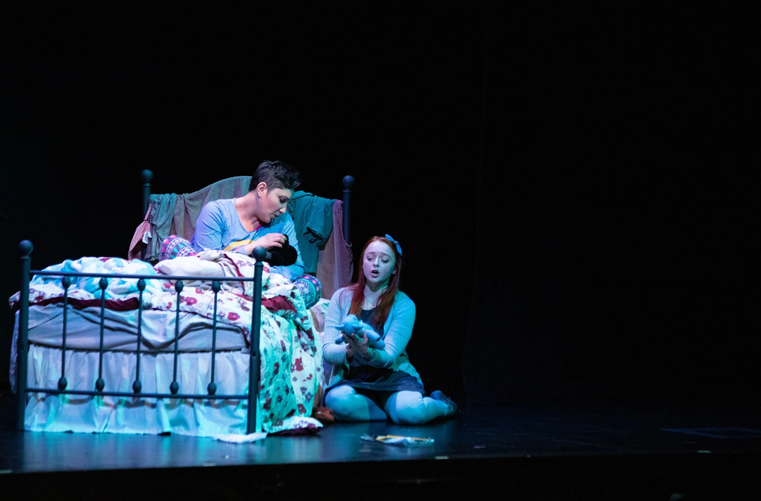 Two young women and a young man are on a stage performing a scene. The young women are sitting with a bed with clothes on it, while the young man is sitting in front of the bed, looking at his phone. The scene is part of an opera.