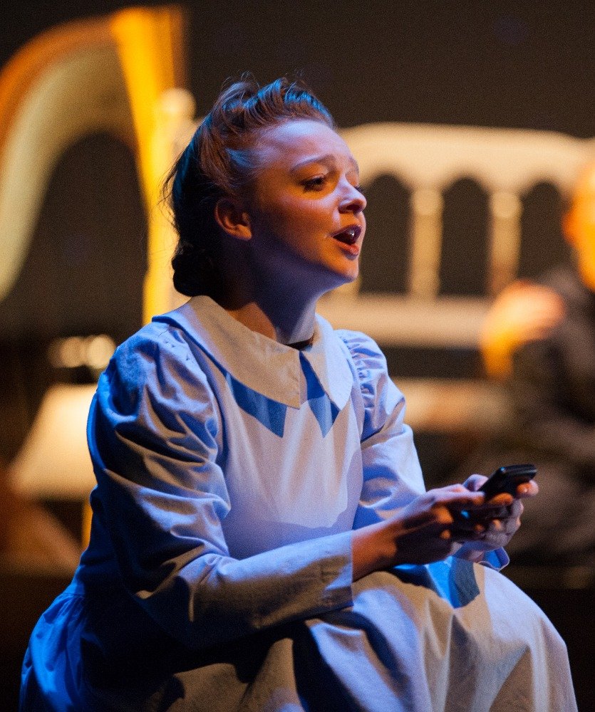 A woman in a vintage-style dress holding a phone, sitting in a dimly lit room, possibly performing or acting.