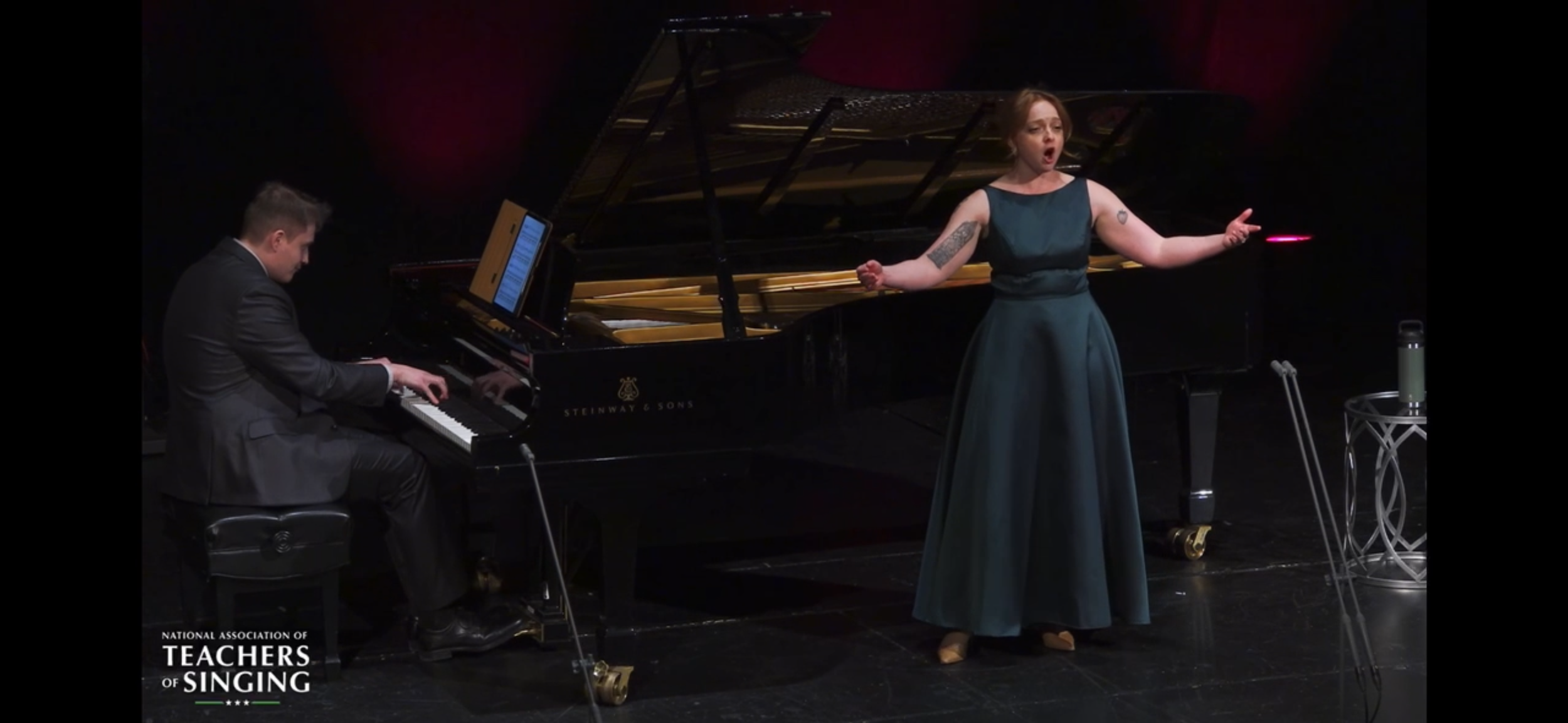 A woman in a teal dress singing on stage with a man playing a grand piano. They are performing at an event organized by the National Association of Teachers of Singing.