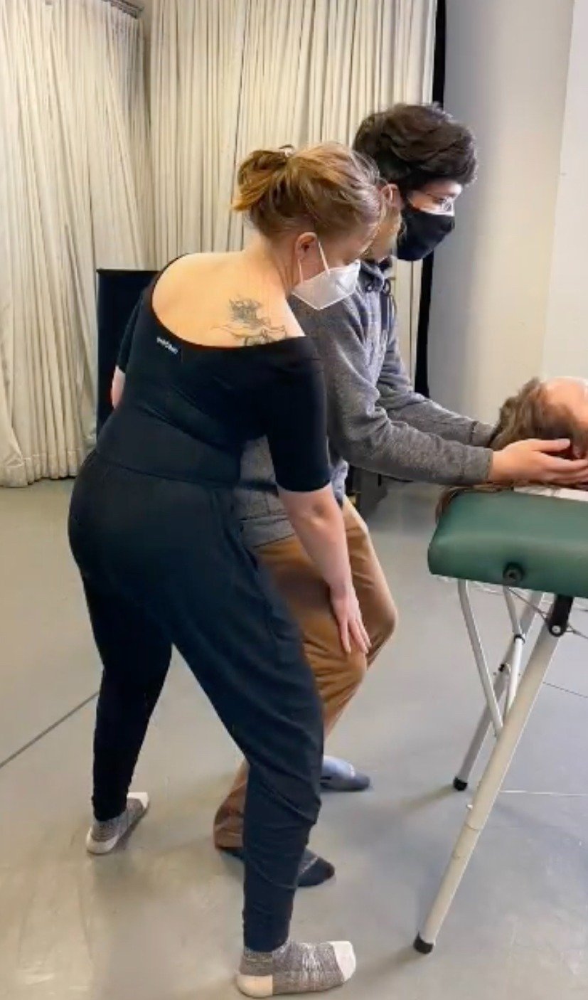 A young woman with red hair and a medical mask adjusts the posture of a young man, also masked, during an Alexander Technique session.
