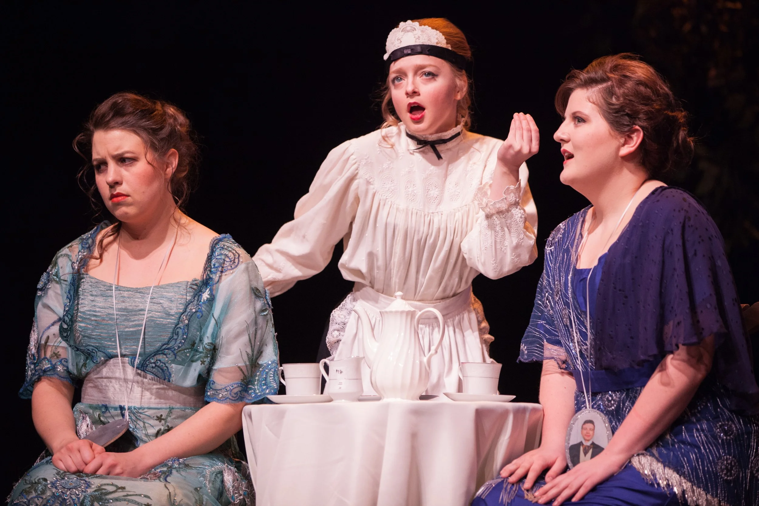 Three women dressed in vintage or period costumes standing around a table with white tableware, engaging in a theatrical scene.
