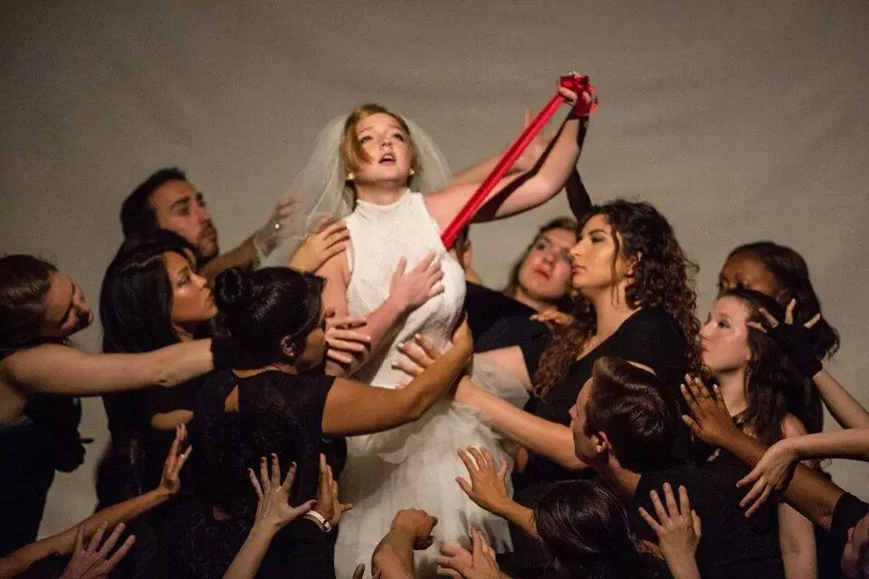 A woman in a wedding dress surrounded by women and men reaching out to her, holding a red ribbon connected to her wrist, in a staged scene.