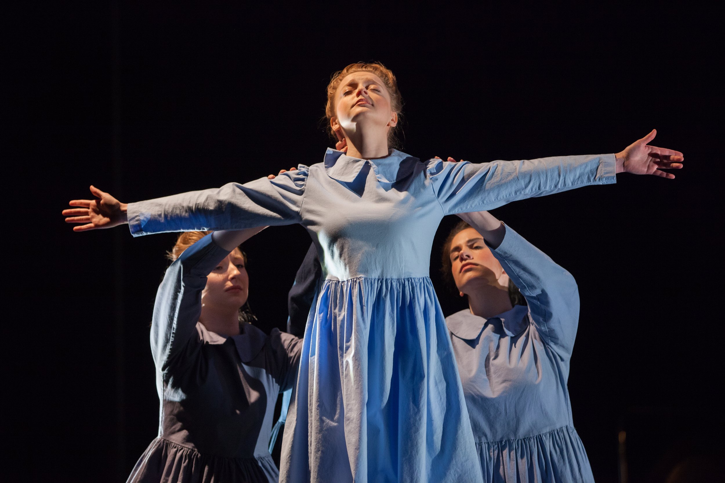 A group of women in matching blue dresses performing a dance or movement routine on stage, with one woman at the center extending her arms outward and eyes closed, against a dark background.