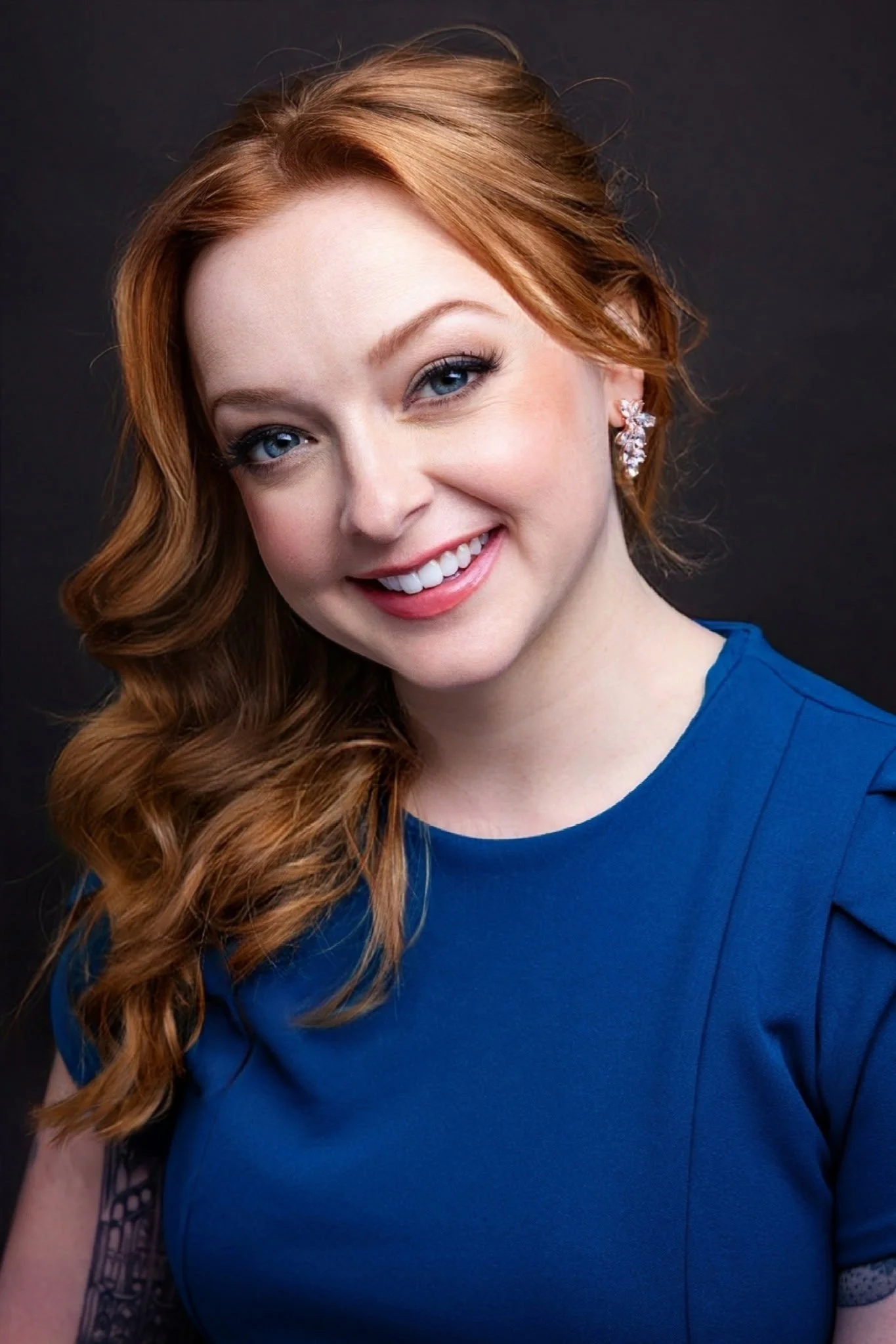 A smiling woman with red hair, blue eyes, wearing a blue top, silver earrings, and a tattoo on her left arm, against a dark background.