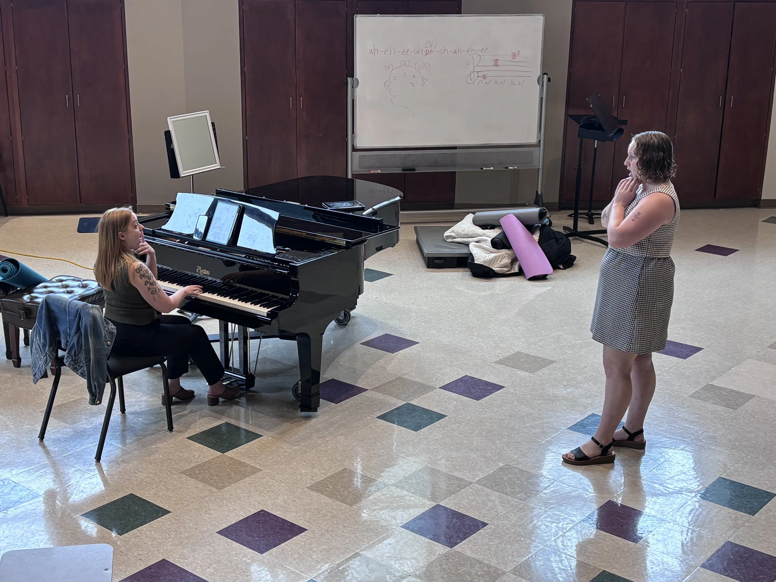 Two women in a room with a grand piano and a whiteboard. One woman is sitting at the piano offering instruction, the other is standing and singing.