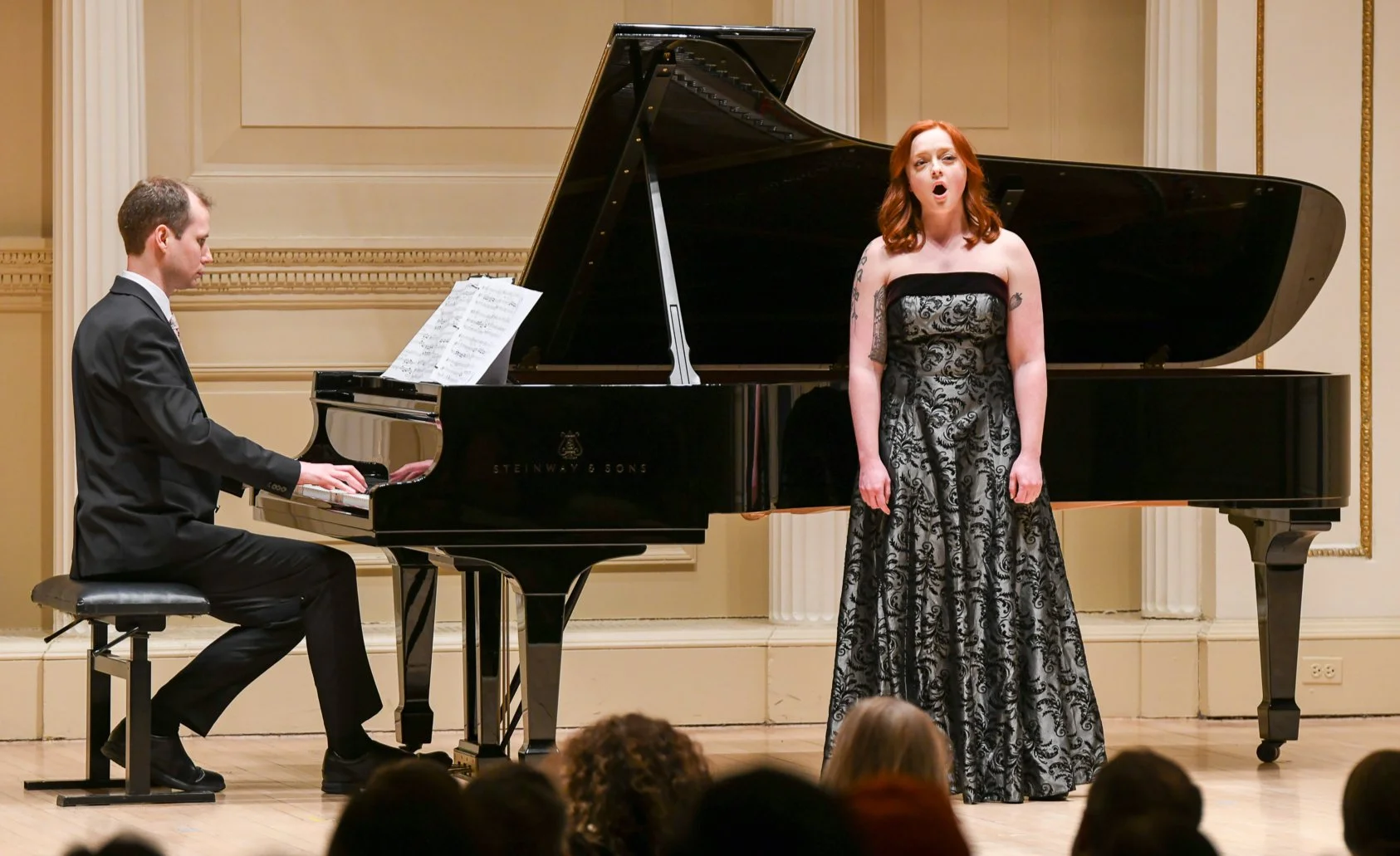 A male pianist playing a grand piano while a female singer stands next to him on stage during a concert in a formal hall.