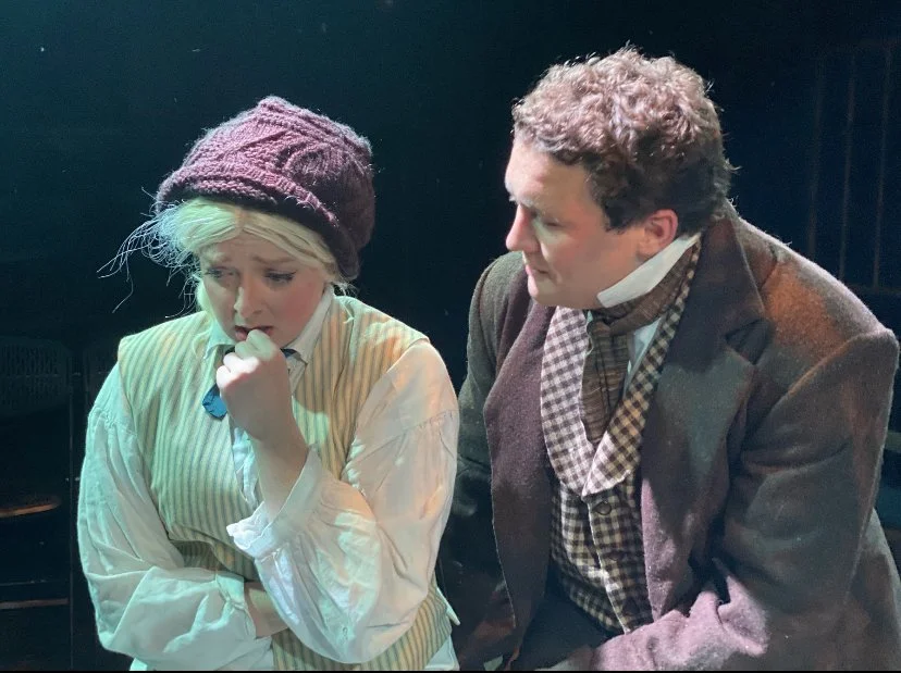 A young woman and an older man dressed in vintage clothing, sitting close together, having a serious conversation.