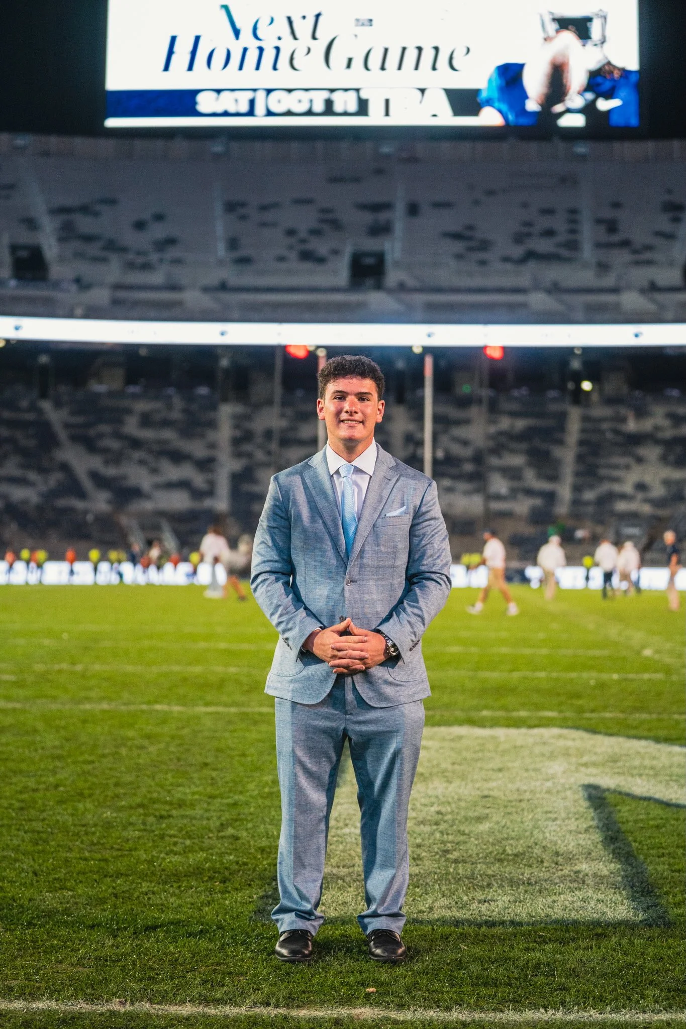 A young man in a light gray suit standing on a football field at night, smiling with hands clasped in front of him, with stadium seats and a scoreboard in the background.