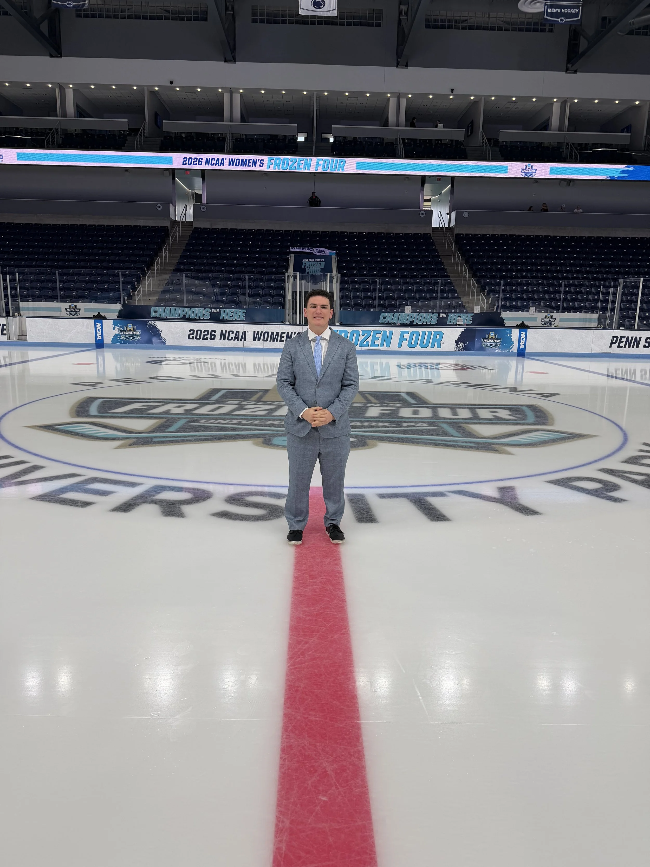 A man in a gray suit standing on an ice hockey rink with a large logo at center ice and digital banners that read '2026 NCAA Women's Frozen Four' and 'Champions Win Here'.