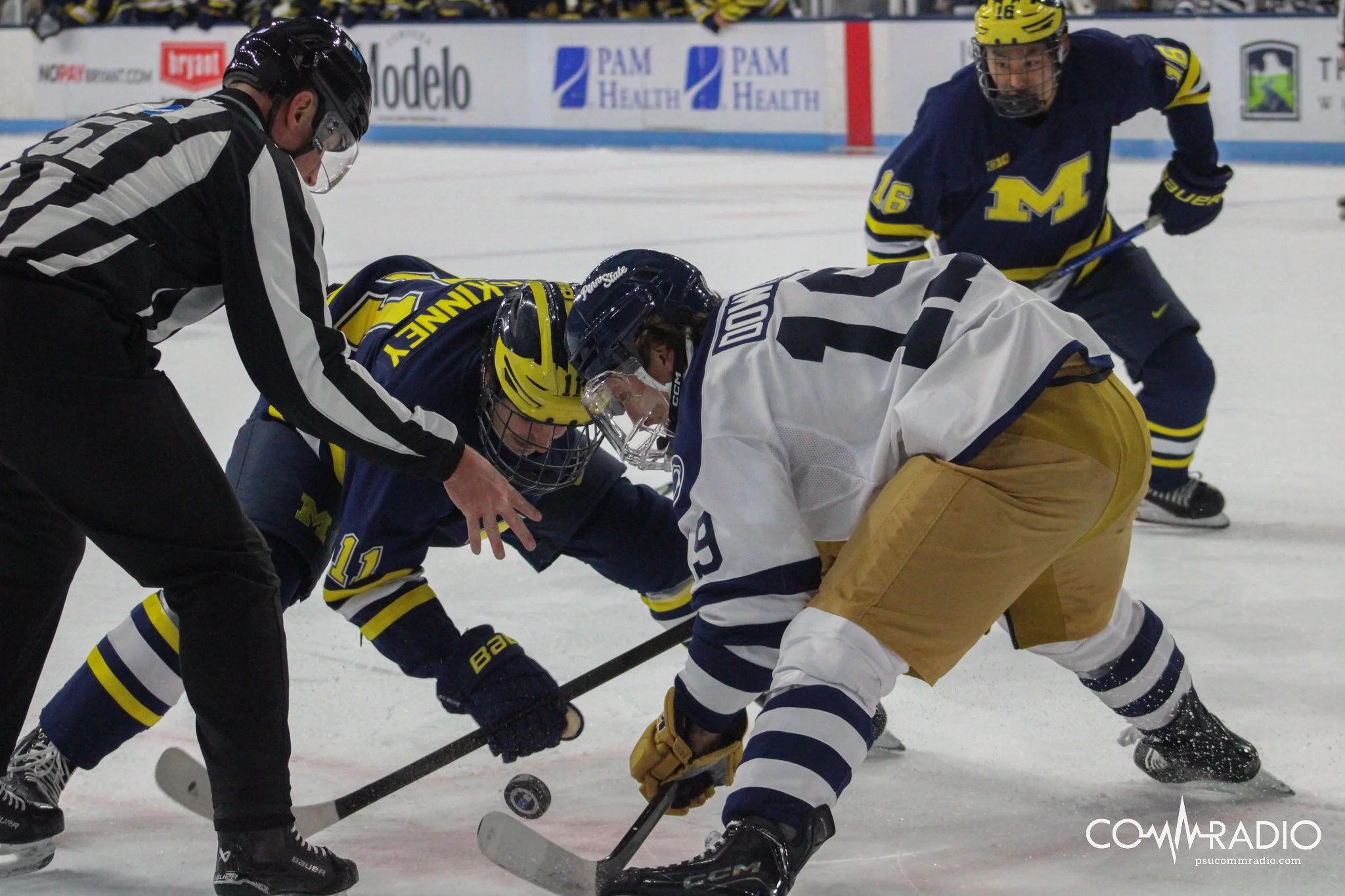 Hockey players are battling for puck control as an official observes during a game on ice rink. One player from Michigan wears a blue jersey with yellow and white accents, while the opponent wears a white jersey with dark blue and yellow accents.