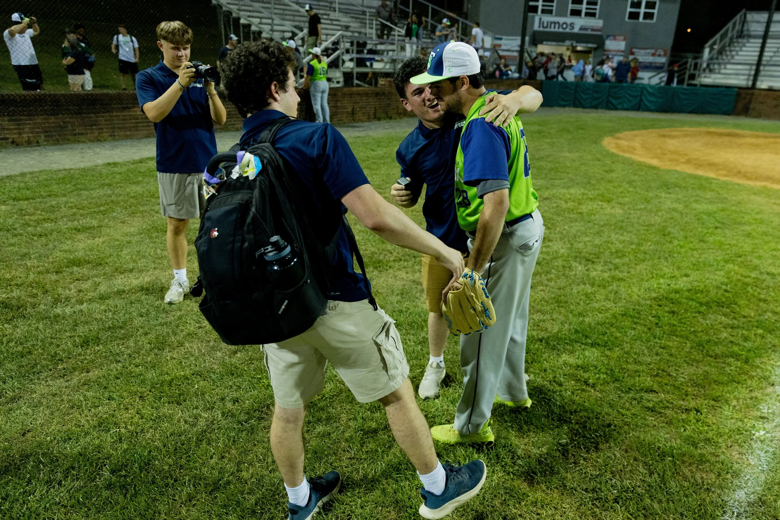 Two men greet each other with a handshake and hug on a baseball field at night, while a boy with a backpack and camera takes a picture nearby and others watch from the bleachers.