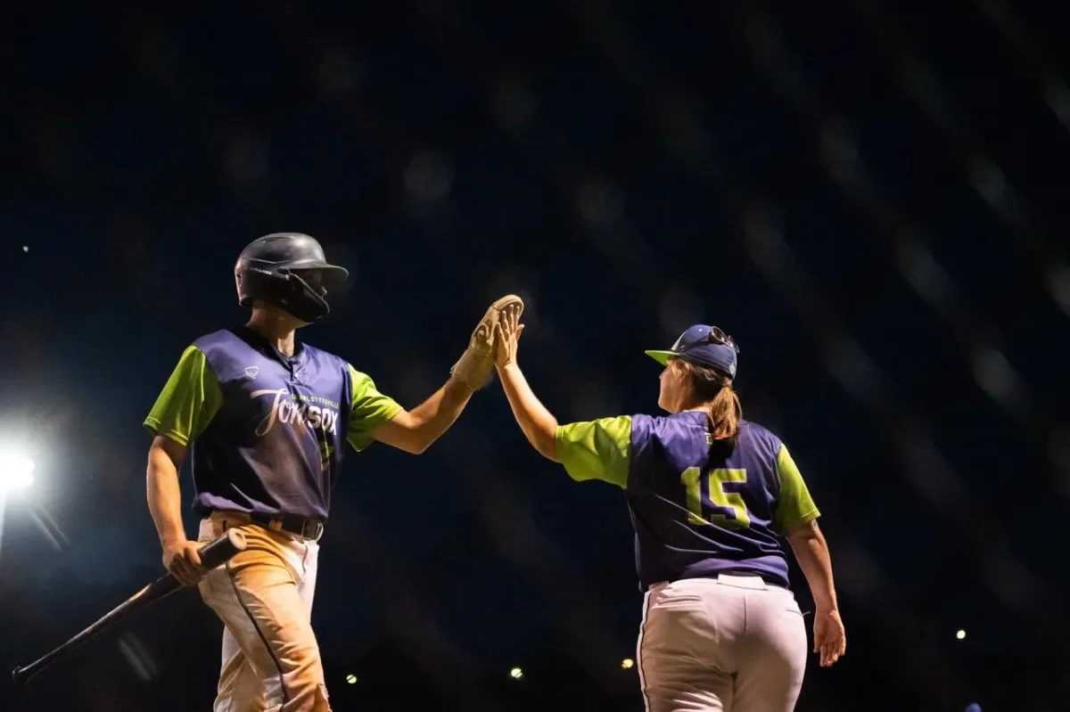 Two female softball players, wearing purple and green uniforms, high-five each other at night.