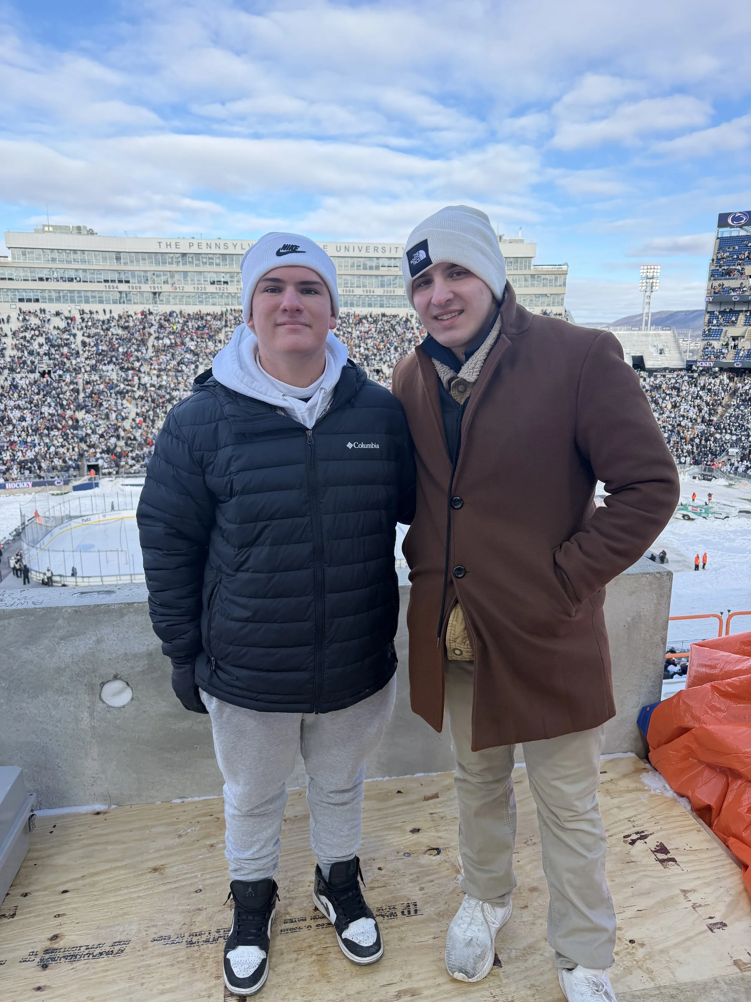 Two young men standing on a wooden platform at a hockey stadium, with the crowd and scoreboard in the background. One is wearing a black puffer jacket and gray sweatpants, the other a brown coat and beige pants, both in warm clothing with beanie hats. The stadium is filled with spectators, and an ice rink can be seen below.