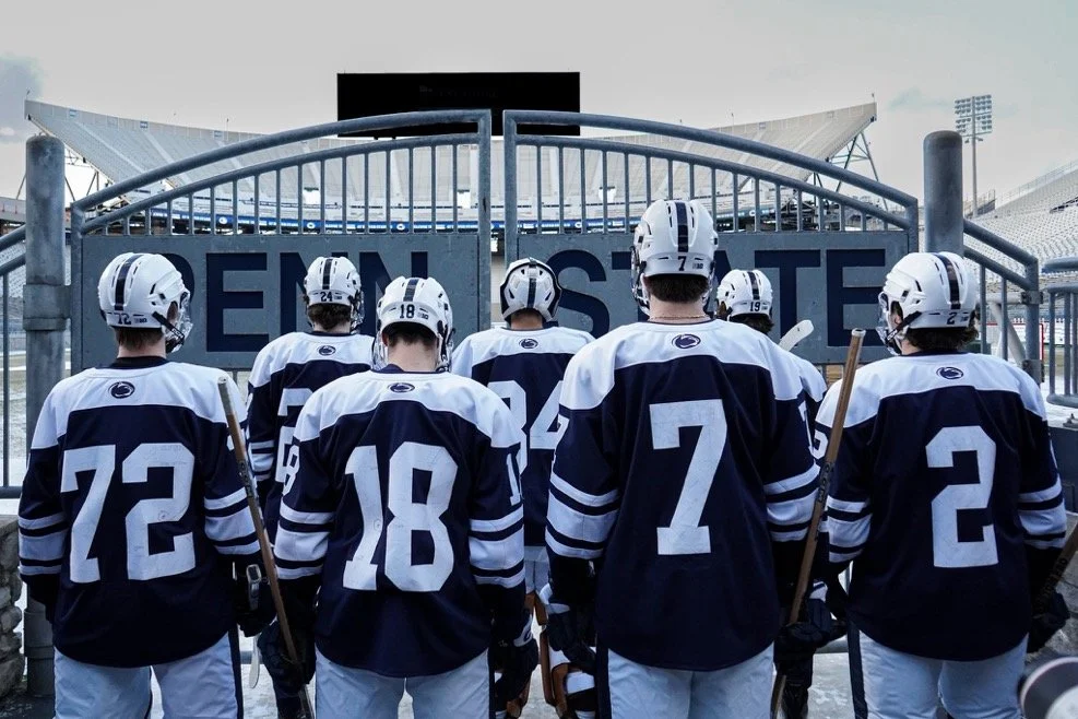 Group of ice hockey players in uniform on the field at a stadium, facing away toward the entrance gate.