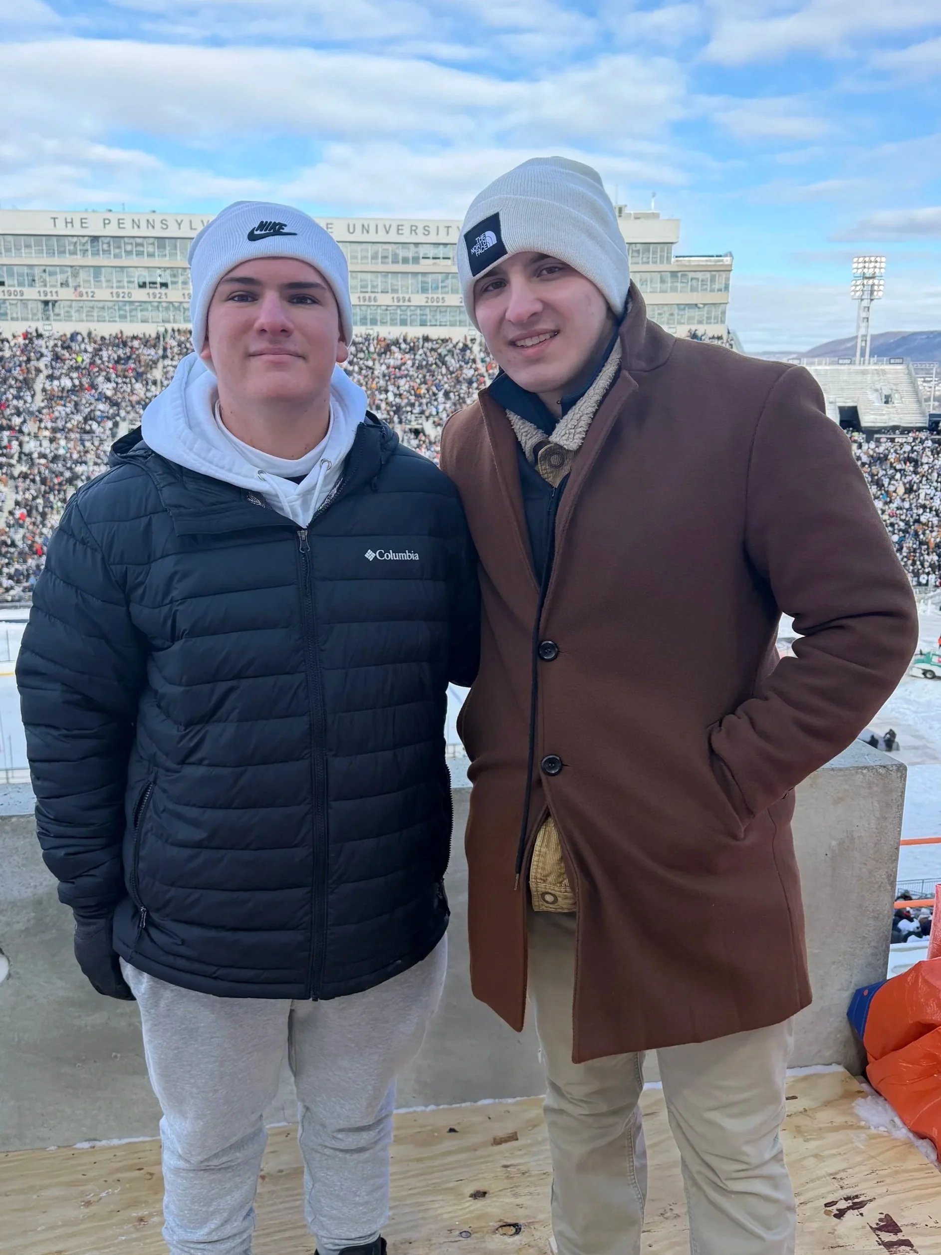 Two young men standing outdoors at a sports stadium, wearing winter clothing and beanies, with a large crowd and a stadium building in the background.