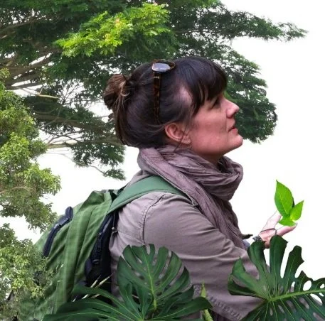 Woman with sunglasses on her head, wearing a backpack and scarf, standing outdoors among green trees and large tropical leaves.