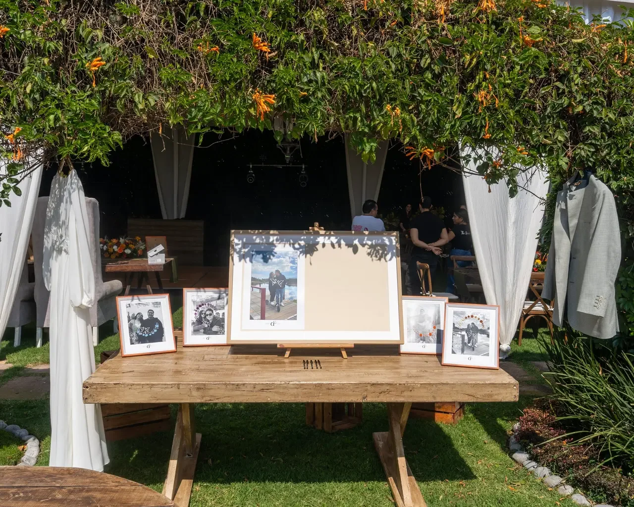 A rustic outdoor wooden table displays framed photographs and a blank canvas, under a leafy archway with white curtains, leading to a gathering of people in a garden setting.