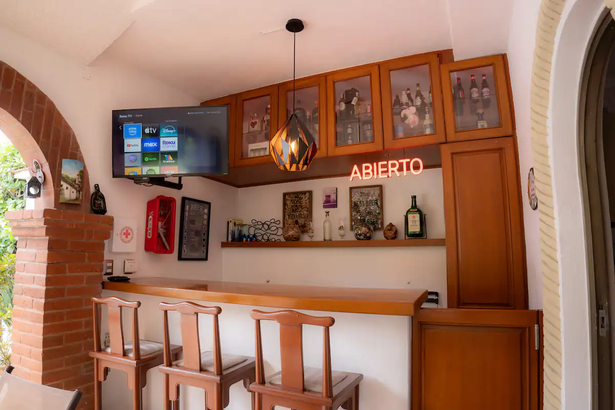 Indoor bar with three wooden chairs at a counter, wooden cabinets and shelves with bottles, a neon sign that says 'ABIERTO,' a television on the wall, and a brick arch with a garden visible outside.