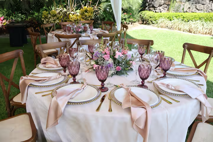 Elegant outdoor wedding reception table set with pink and gold accents, floral centerpiece, pink glassware, white plates, gold cutlery, and pink napkins, surrounded by wooden chairs in a green garden setting.