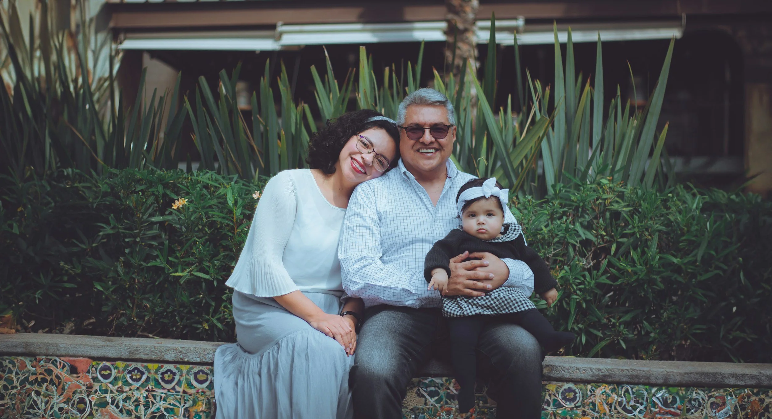 A family of three sitting on a colorful mosaic bench in a garden. The woman has curly dark hair, glasses, and is wearing a white dress. The man has gray hair, sunglasses, and is wearing a checkered shirt. The young girl with a white headband and black sweater is sitting on the man's lap.