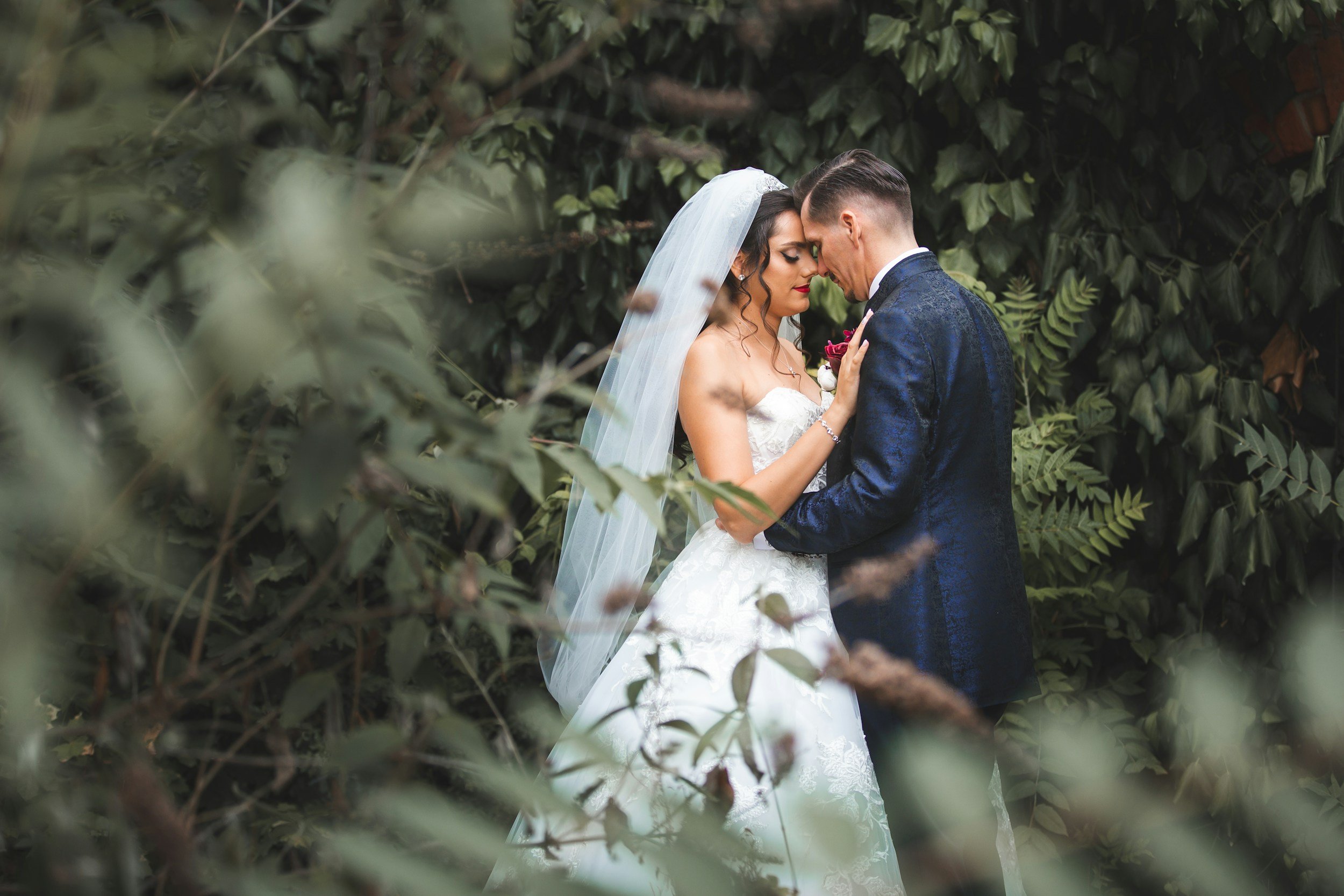 A bride and groom share a close moment with foreheads touching, surrounded by greenery.
