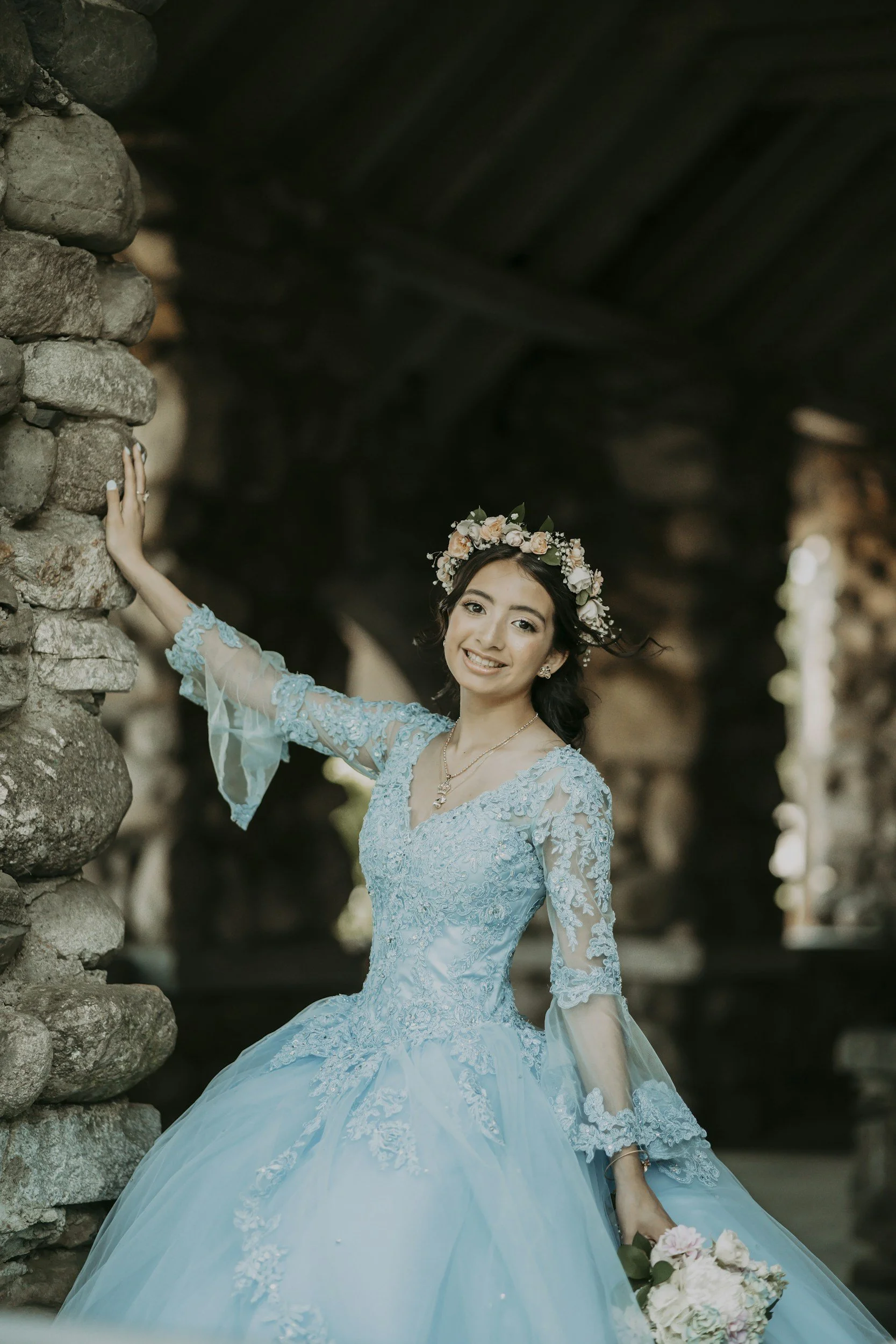 A smiling woman in a light blue, floral embroidered wedding dress with lace sleeves, wearing a flower crown, holding a bouquet of white and pink roses, and standing beside a stone wall.