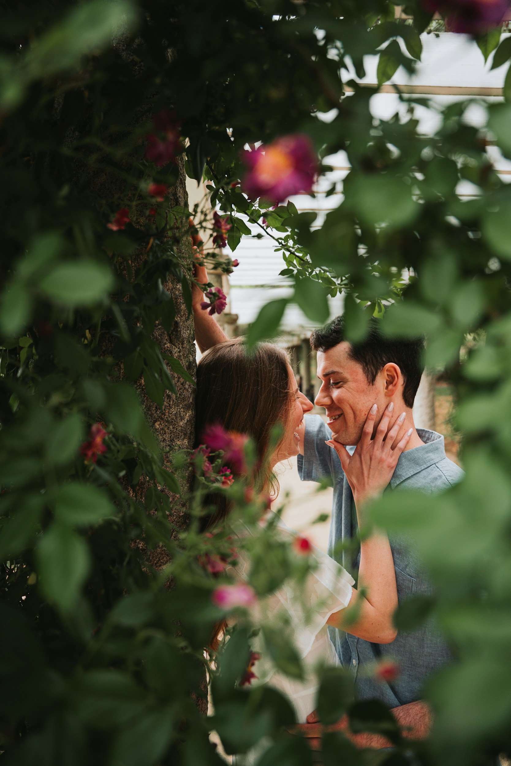 A couple is sharing a romantic moment outdoors, surrounded by green foliage and pink flowers, with their faces close together.