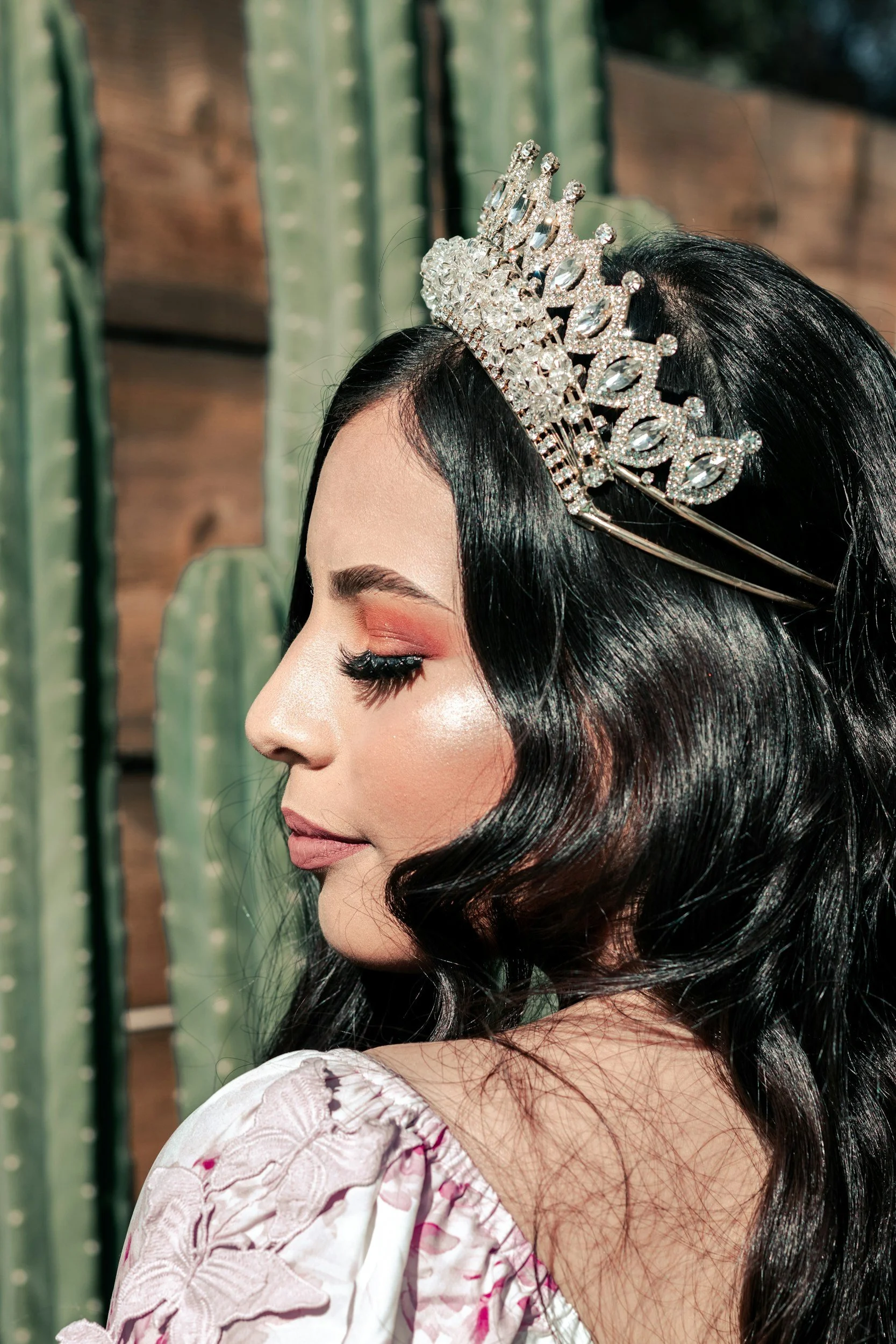 Close-up profile of a woman with dark wavy hair wearing a silver tiara with rhinestones, against a backdrop of wooden panels and cactus plants.