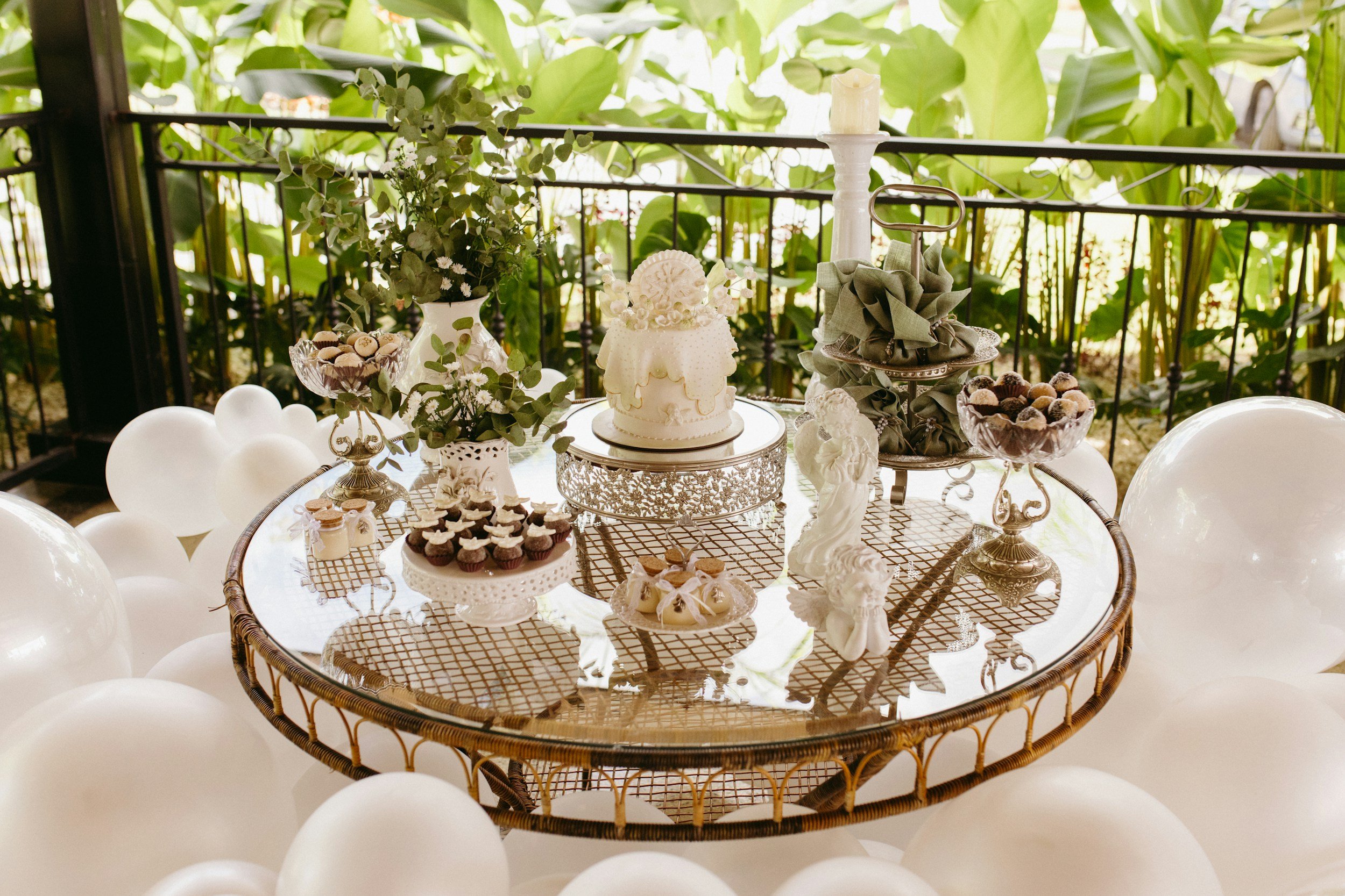 Decorative table with a variety of desserts including cupcakes, chocolates, and cookies, arranged with greenery and a white cake, set on a glass-topped wicker table surrounded by white balloons, outdoors with lush green foliage in the background.