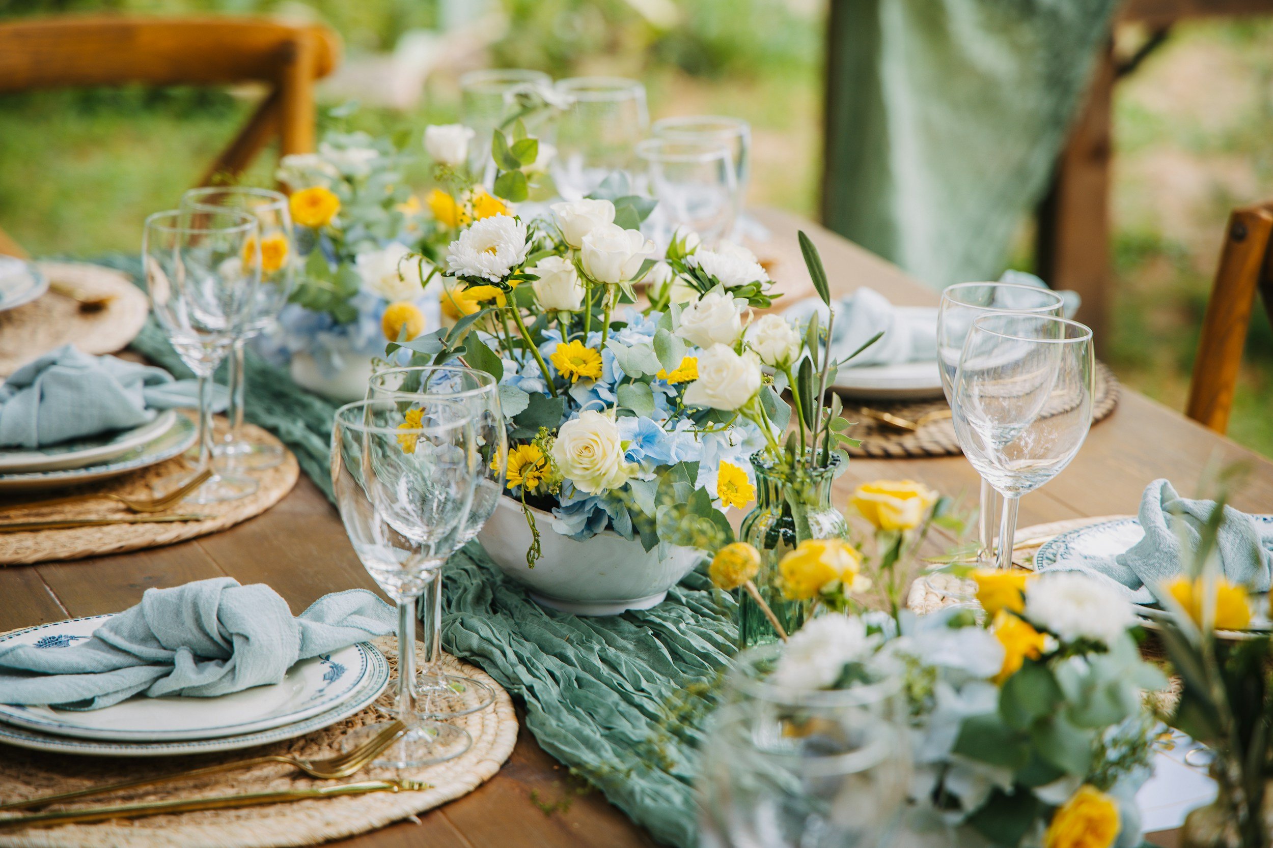 A beautifully set outdoor dining table with a floral centerpiece featuring white and yellow flowers, surrounded by clear wine glasses, plates, and light blue cloth napkins, with a lush garden background.