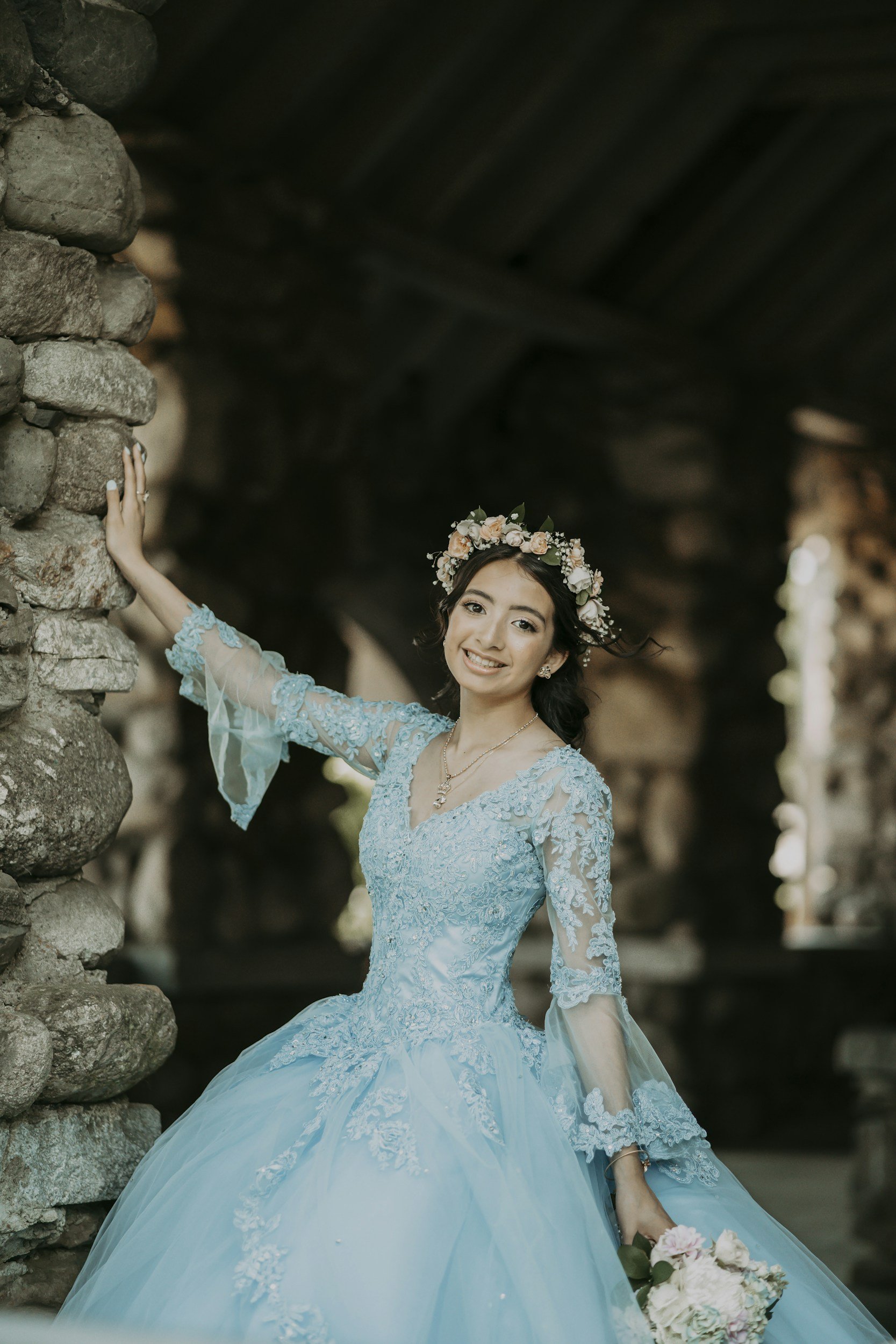 A young woman wearing a light blue, embroidered, long-sleeved ball gown with a full skirt, holding a bouquet of flowers and smiling at the camera. She has a floral crown on her head and is standing near a stone wall in a shaded outdoor setting.