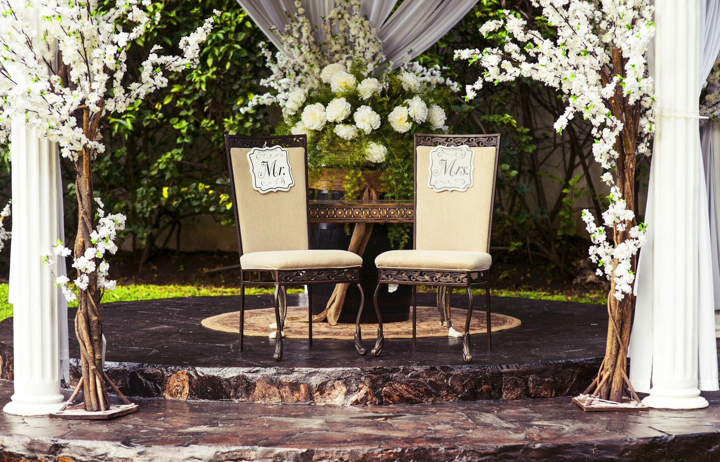 A wedding or event arch decorated with white flowers and white drapes, with two chairs labeled 'Mr.' and 'Mrs.' in front, set on a stage with a tree trunk and greenery in the background.