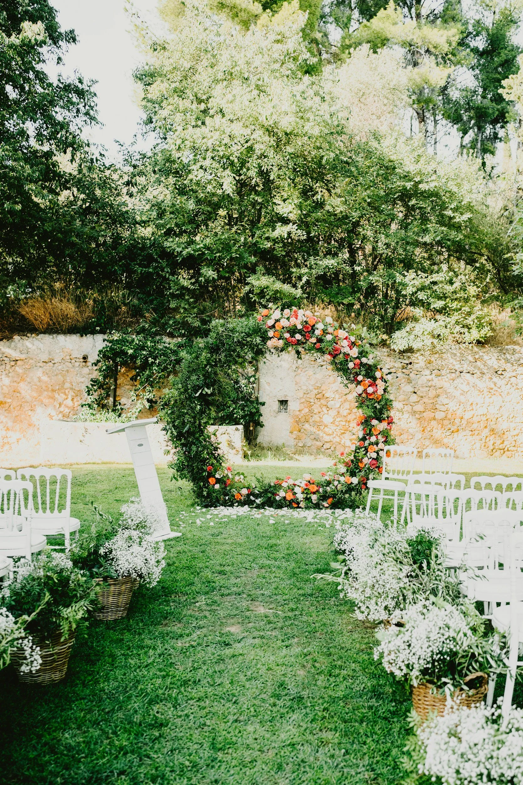 Outdoor wedding ceremony setup with white chairs, flower arrangements in baskets, and a circular floral arch on a grassy lawn surrounded by trees.