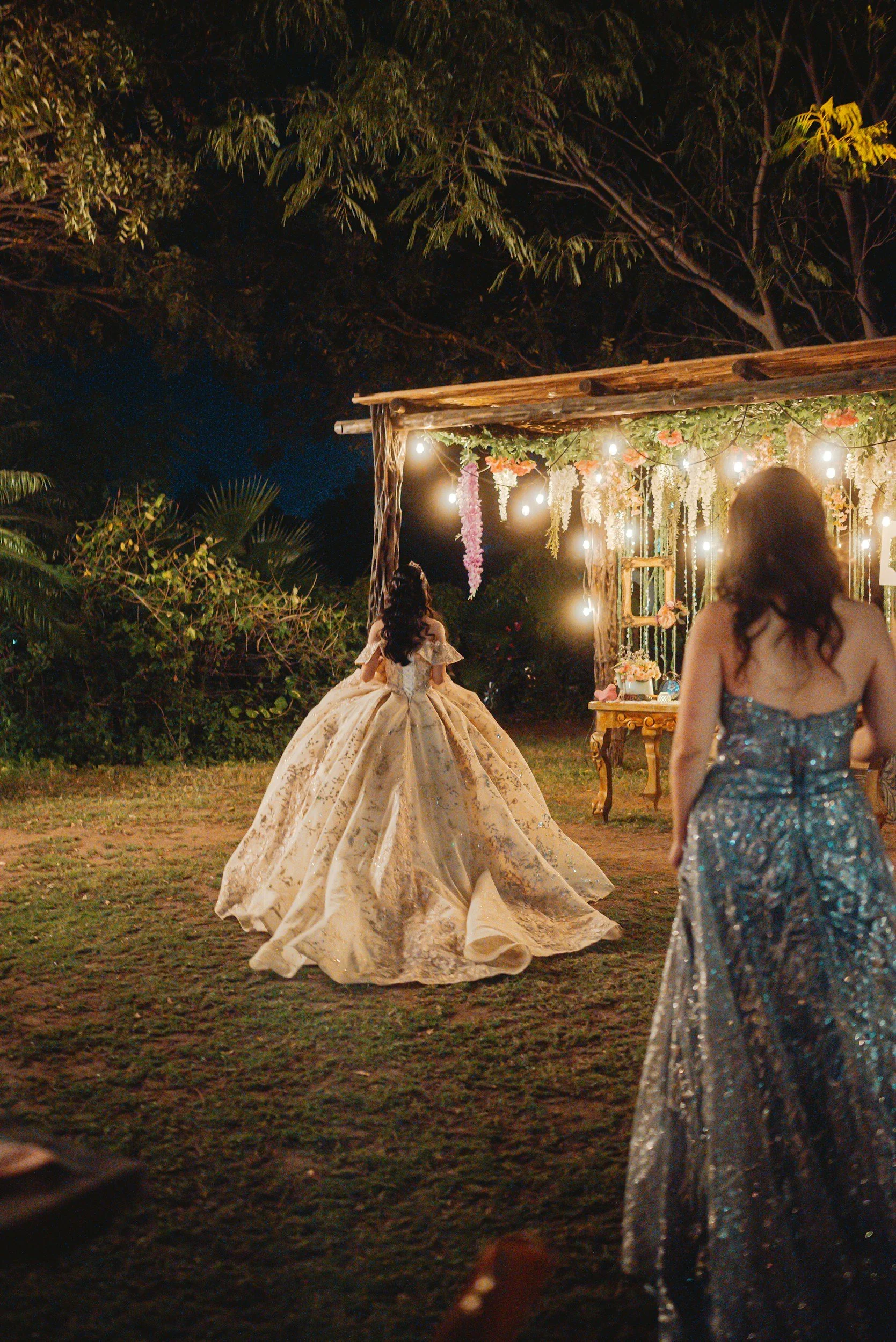 Nighttime outdoor event with two women in elegant dresses, one in a ball gown, and string lights with hanging flowers on a wooden structure, surrounded by trees.