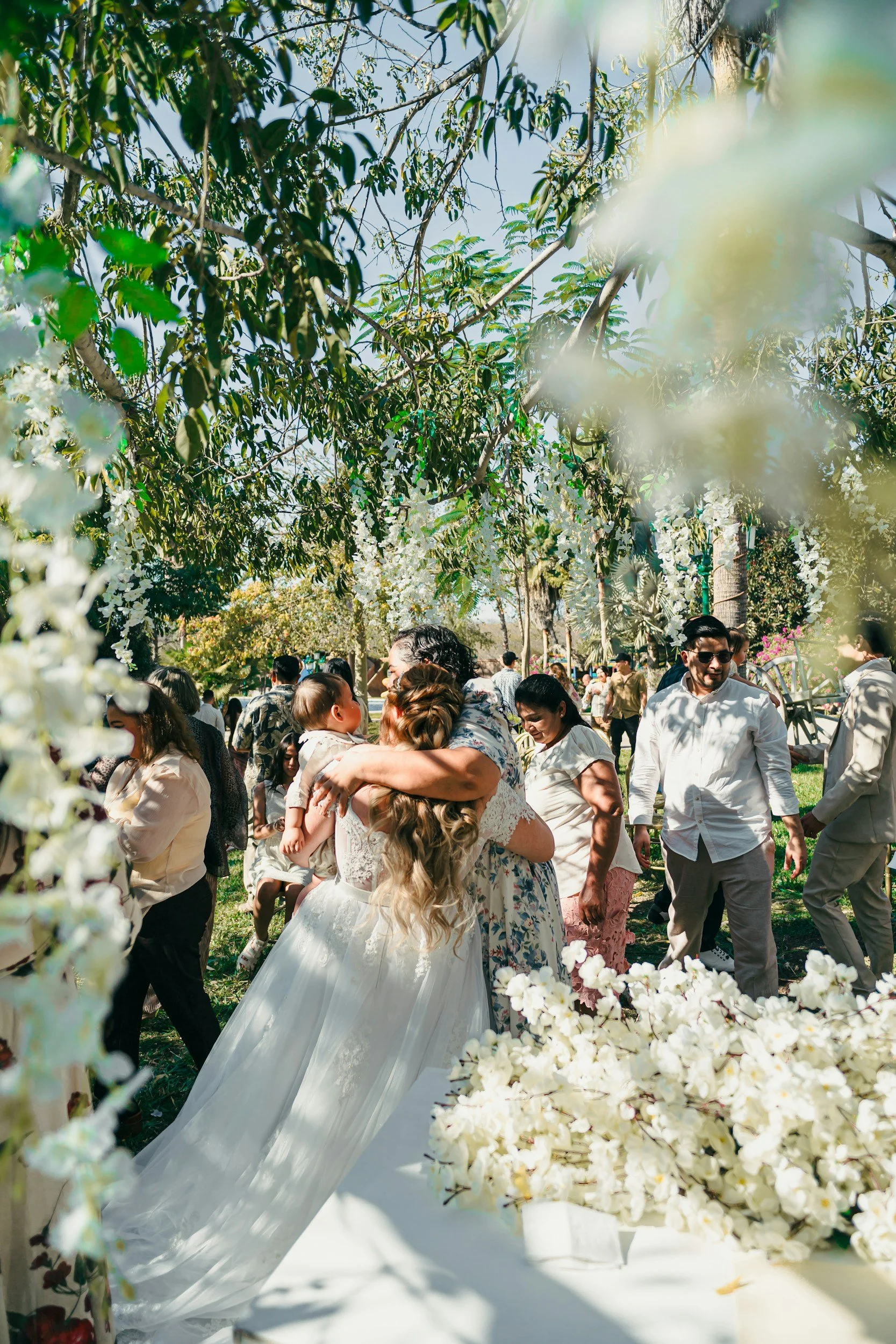 A bride in a white wedding dress with long blonde hair hugs a woman in a floral dress, surrounded by guests at an outdoor wedding ceremony decorated with white flowers and green foliage. The scene takes place amidst tall trees on a sunny day.
