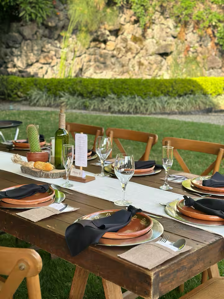 Outdoor dining table set with plates, black napkins, wine glasses, and a wine bottle, with a garden and rocky background.