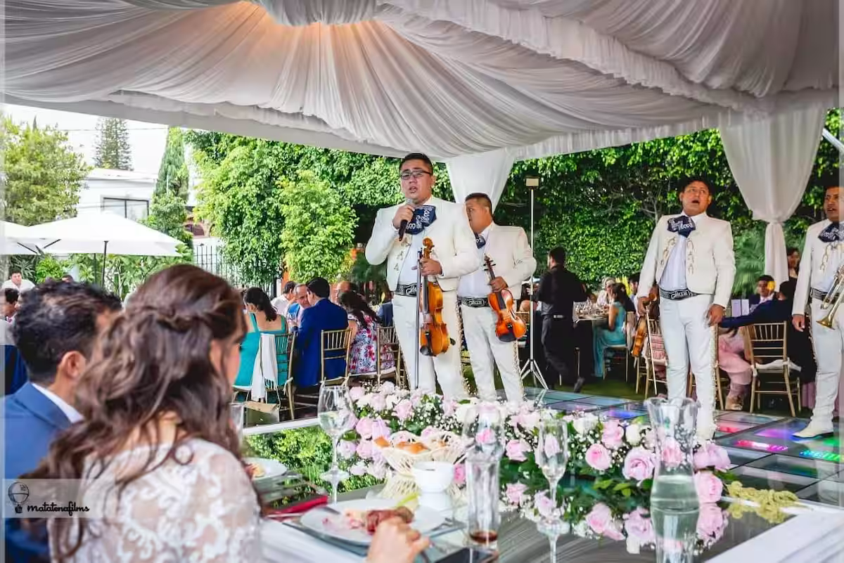 A live band performing at a wedding reception under a large white tent with floral decorations, surrounded by seated guests at tables outdoors.