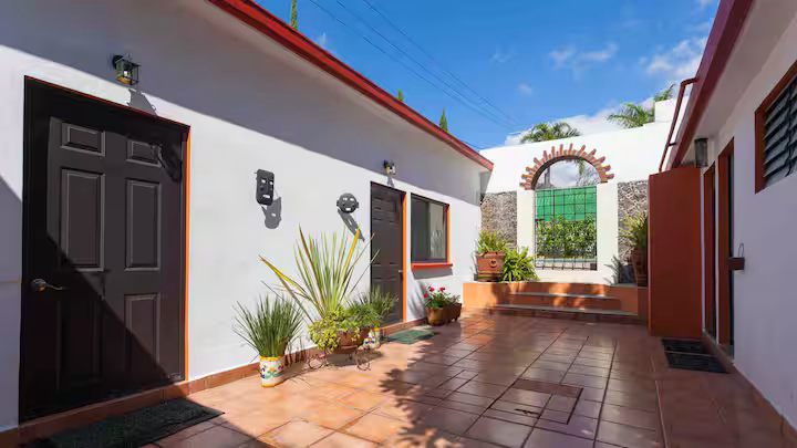 A sunny courtyard with white walls, terracotta tiled floor, and potted plants, including a cactus and flowers. There are doors and windows on the walls, with a decorative brick arch and green gate at the far end.